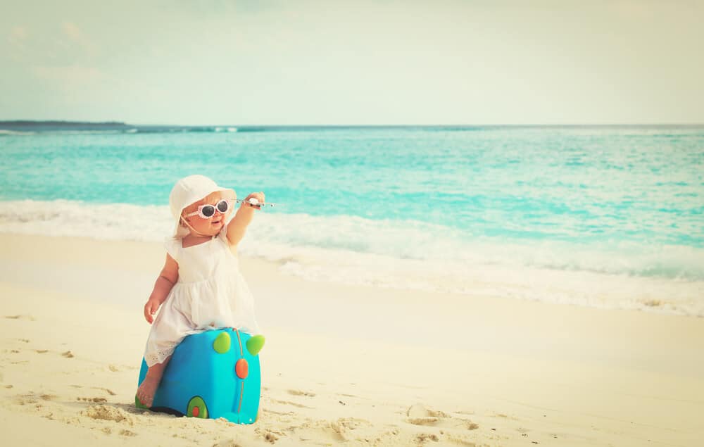 Reisen mit Babys - Kleines Mädchen sitzt auf einem Koffer am Strand